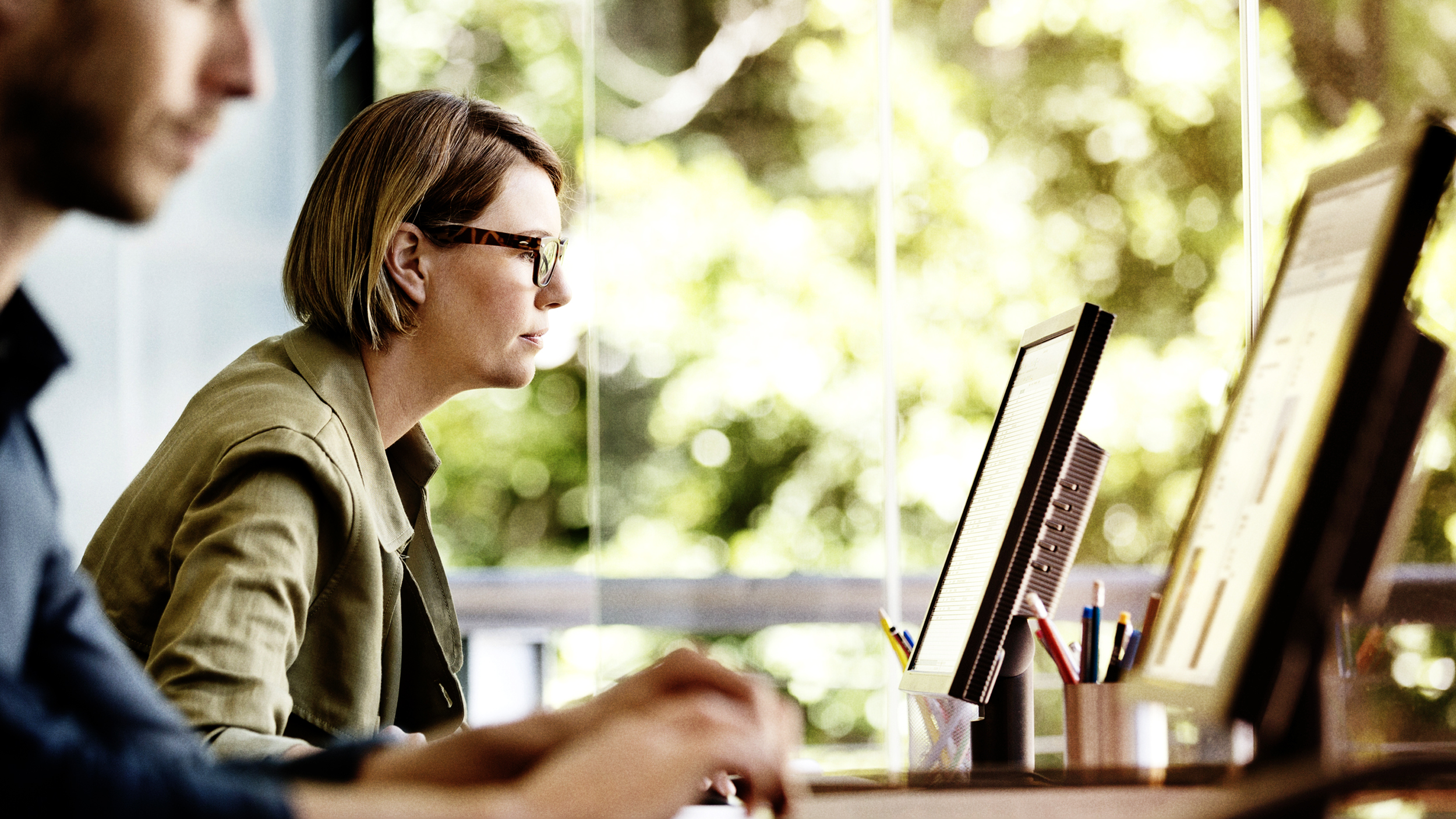 Two IWF Analysts working at computers, looking at screens with a window in the background looking out onto green plants