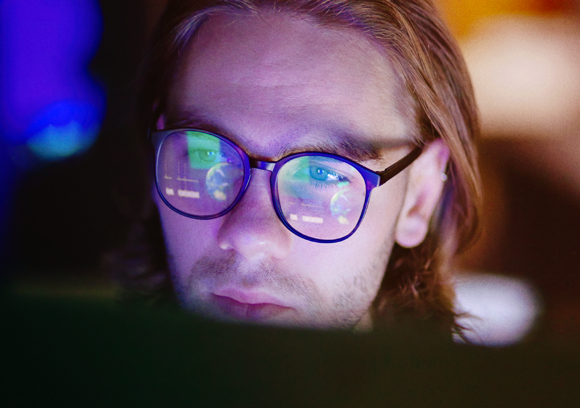 Man wearing glasses sitting behind a screen in a dark room, the image reflecting on the glass of his glasses