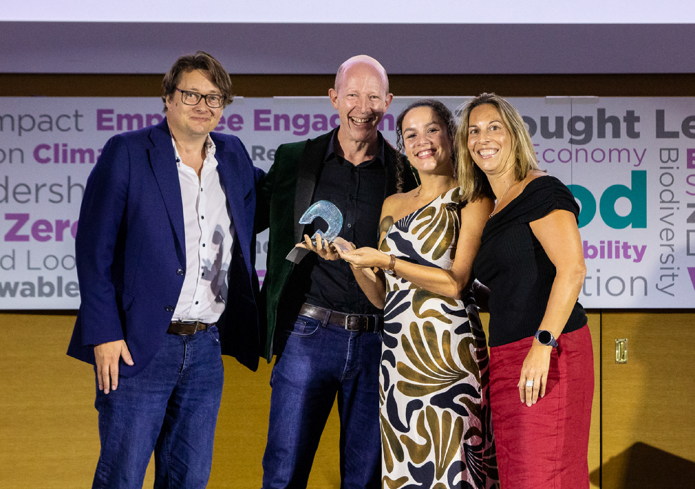 Four people smiling on stage at the Global Good Awards, holding a blue glass trophy, in front of a backdrop of sustainability-themed words.