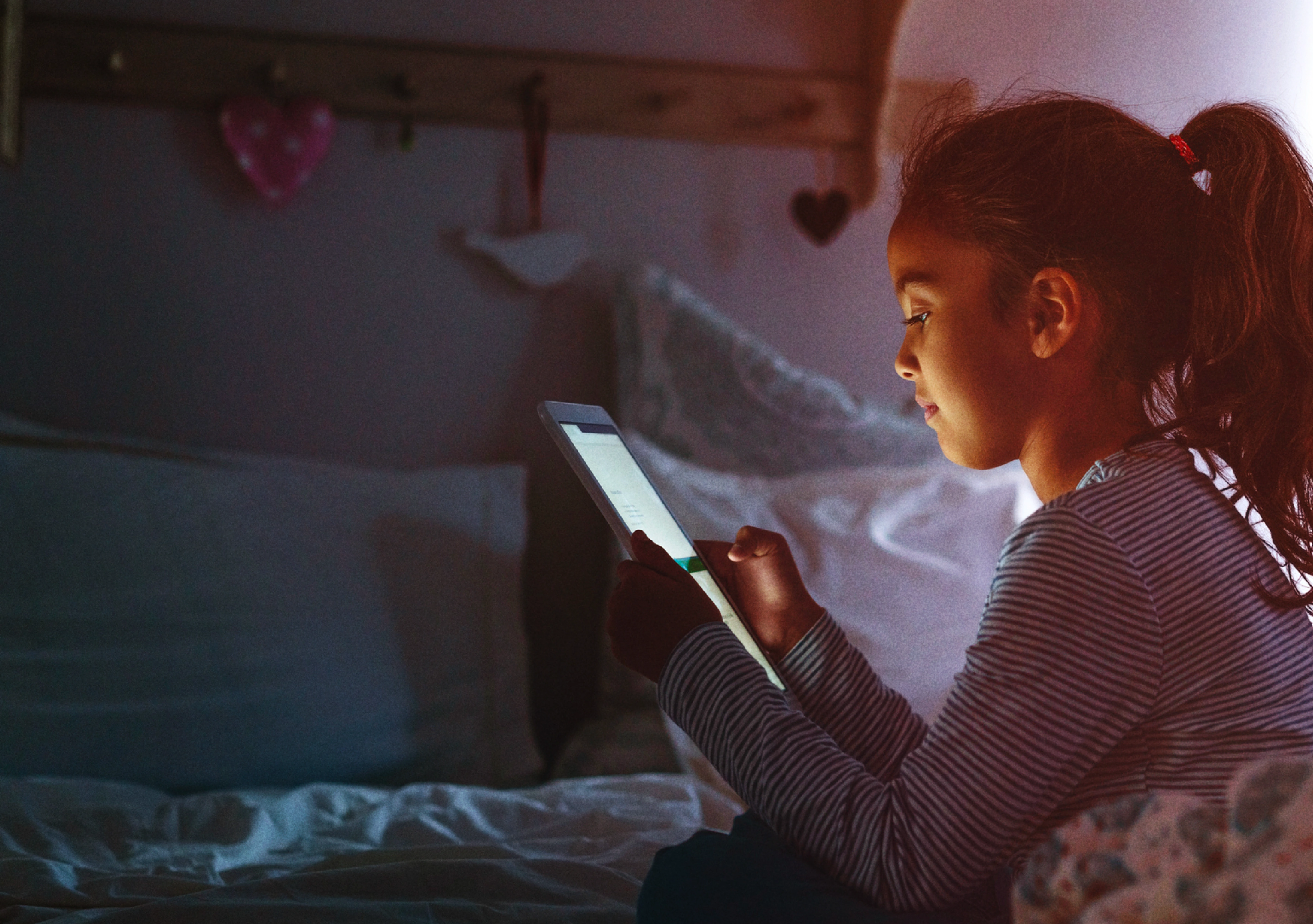A young girl sits in her bedroom, illuminated by the glow of a bright tablet screen reflecting on her face.
