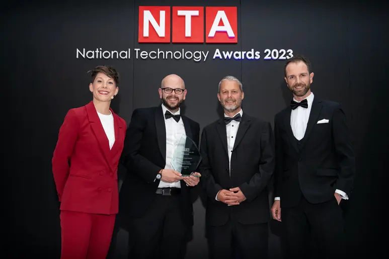Four people wearing smart clothes standing infront of the National Technology Awards logo holding an award