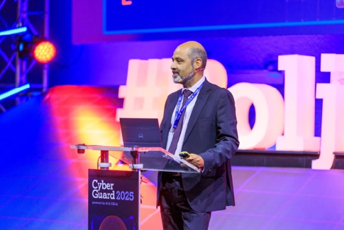 IWF Hotline Director Chris Hughes speaks on stage at the Cyber Guard 2025 conference, standing at a clear podium with a laptop and conference branding visible, illuminated by colourful stage lighting.