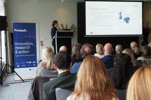 An audience sits in a conference room watching a presenter speak at a podium beside a large screen displaying a slide about the Protech consortium. A branded Protech banner stands to the left of the stage.